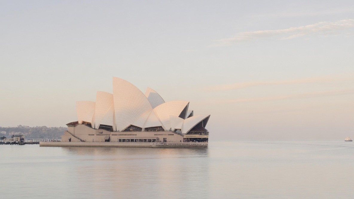 The Sydney Opera House across still harbour water in early morning light.